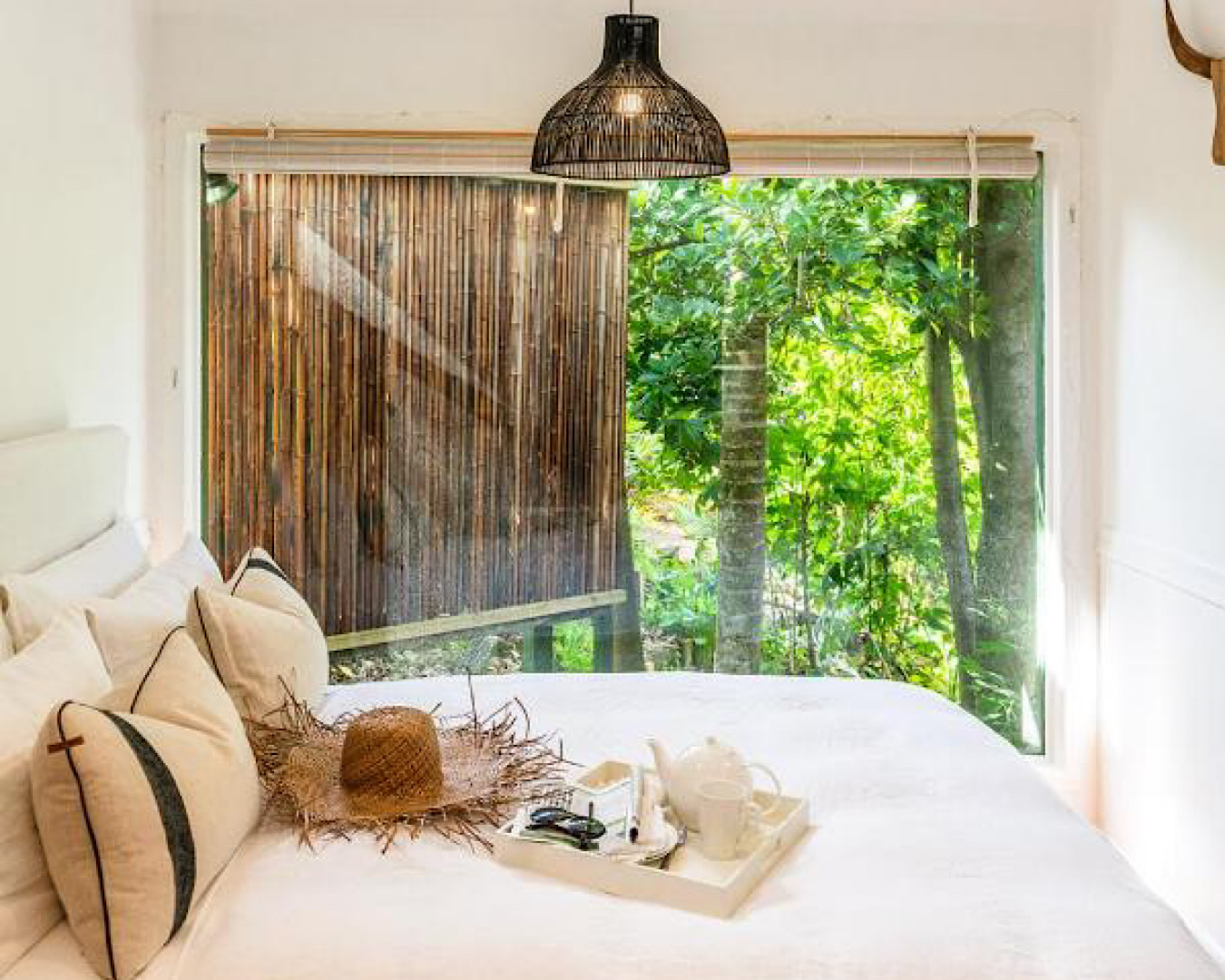 A large, cosy bed is dressed in white with a straw hat and tray with teapot and mug sitting on it. The far wall appears made of bamboo and opens out onto lush greenery. 