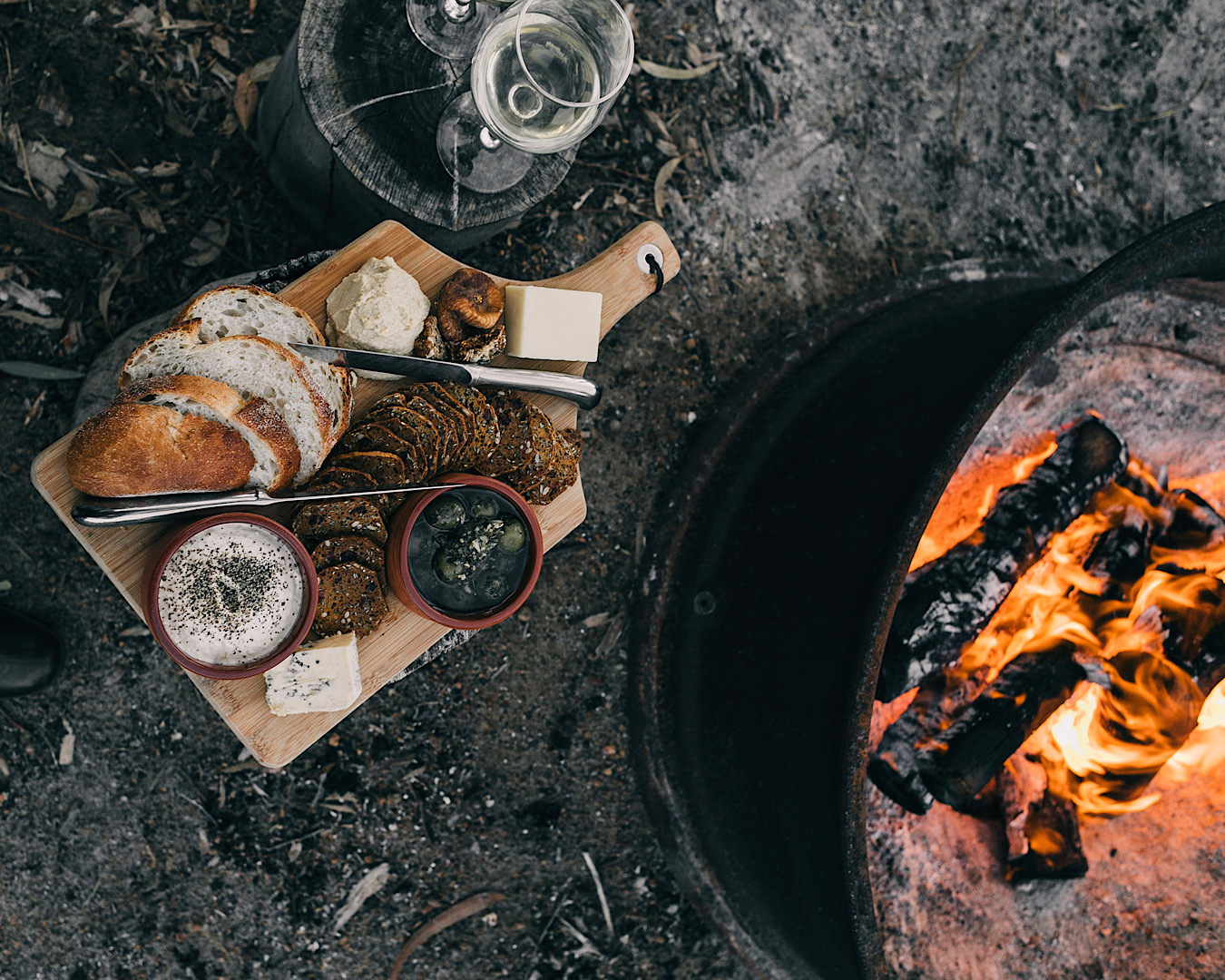 A swanky platter of crackers, dip, olives, bread and cheese sits beside a fire in an oil drum. 