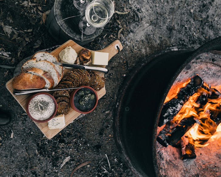 A swanky platter of crackers, dip, olives, bread and cheese sits beside a fire in an oil drum. 