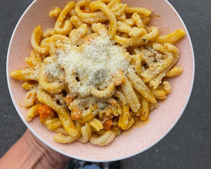 A pink bowl of pasta swirls topped with cheese and veges. 