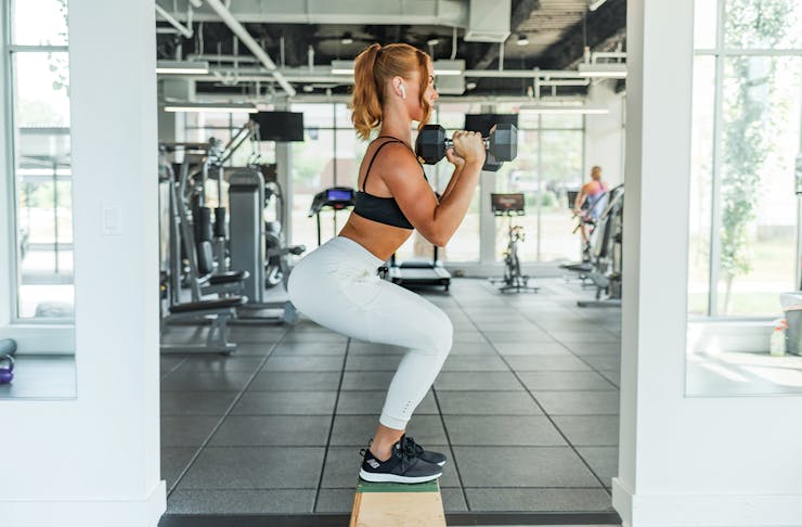 A woman in workout gear squats on a step in a gym.