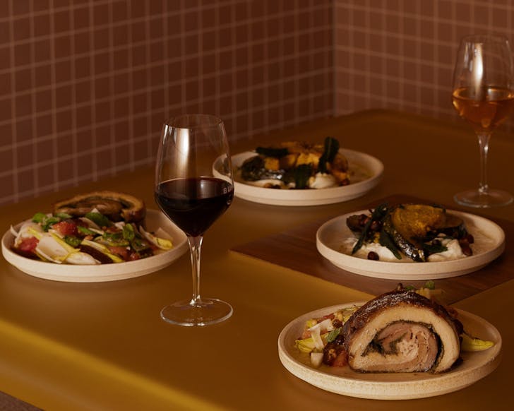 Dimly lit table setting topped with crispy porchetta, salad, roasted pumpkin and glasses of red wine.