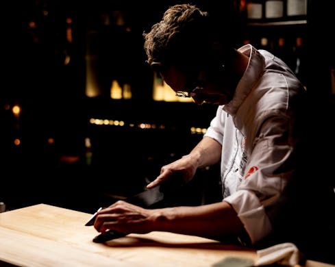A single chef is pictured partly in shadow, using a knife to cut fish on a chopping board