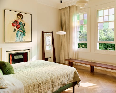 A bed in a light filled room with stained glass windows and a large painting of a woman