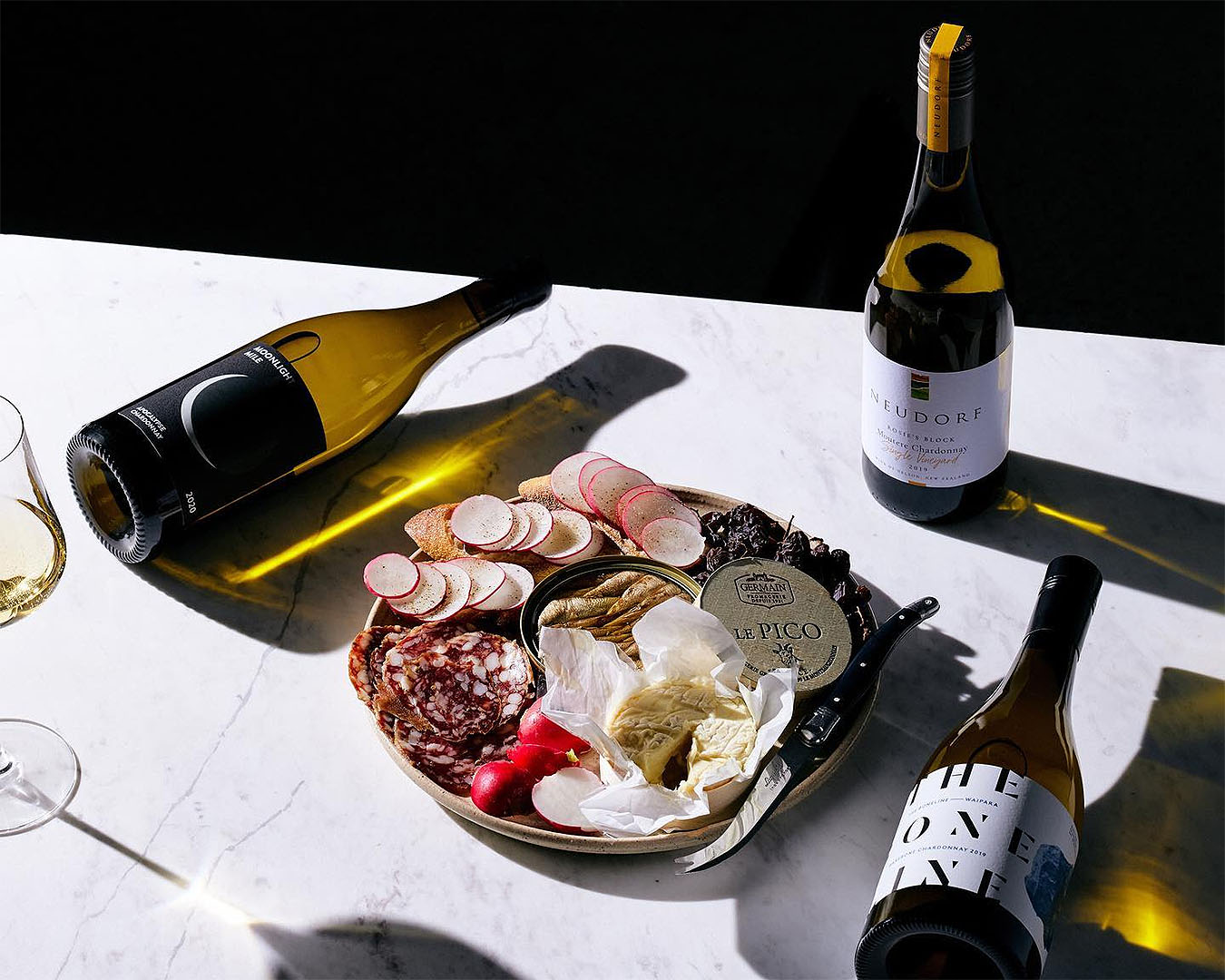 Three chardonnays lie artfully on a table around a platter of food. One of the best alcohol for delivery companies in NZ.