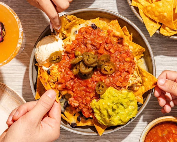 An above view of a large plate of nachos on a dinner table, with little corners of other dishes in the image. Three hands from different angles pick a nacho from the large, main dish.