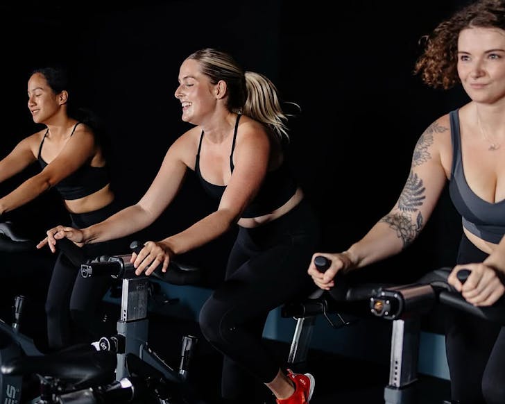 Three women smile and ride it out on stationary bikes. 