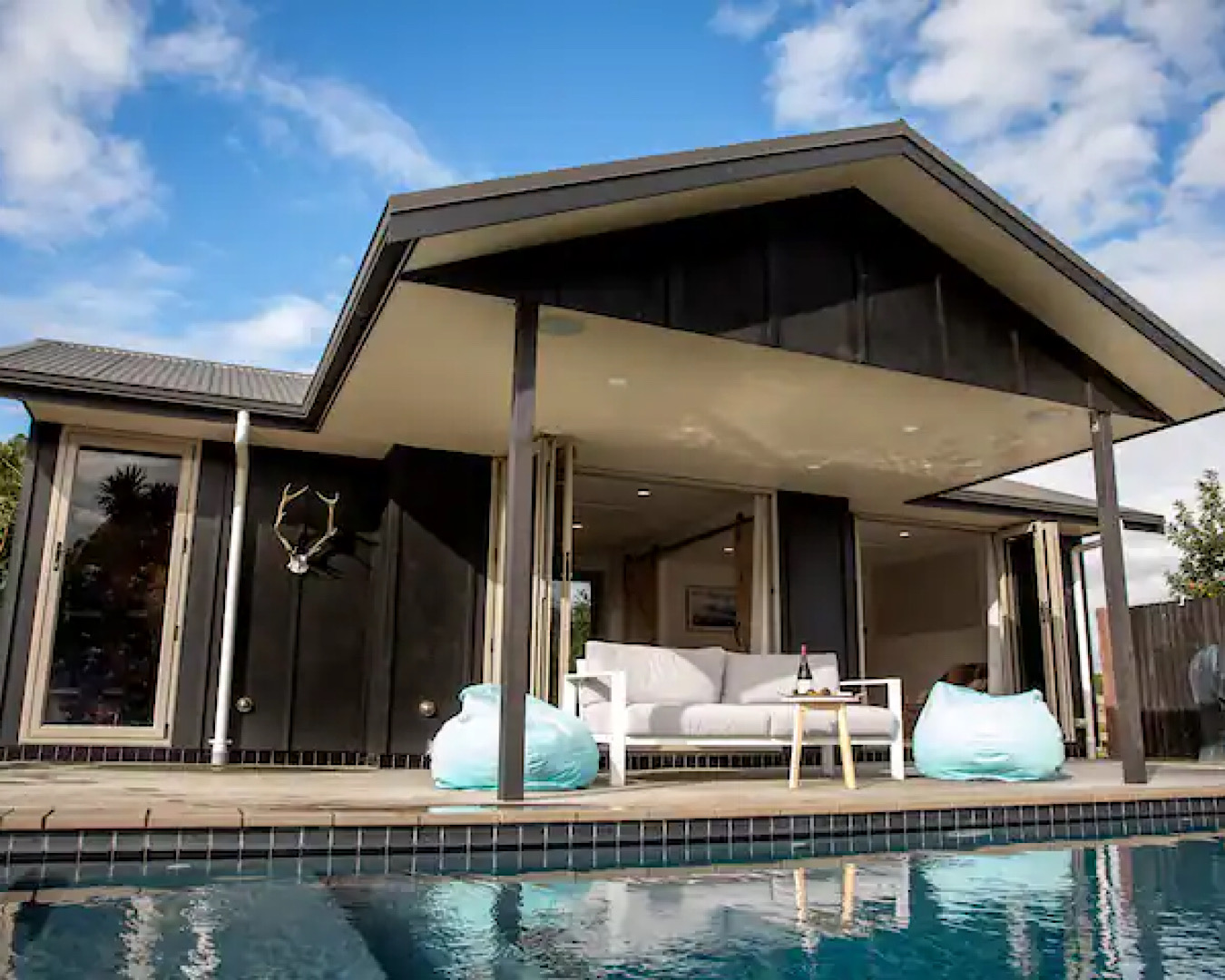 A couch and beanbags with a small table and bottle of wine overlook a pool at the Cambridge Pool House airbnb. 