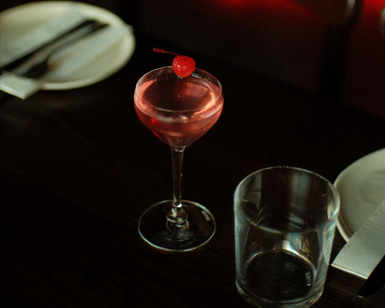 A dark space almost lit by an understated pink cocktail perfectly accented by a maraschino cherry.