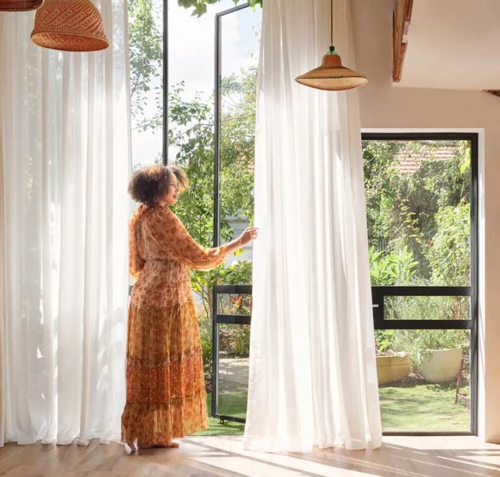 A woman stands by a window with sheer curtains from Carpet Court