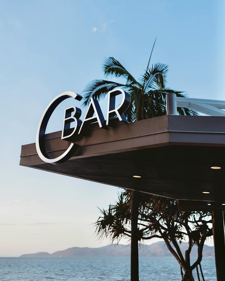 The CBar sign against a backdrop of swaying palm trees and the Coral Sea.