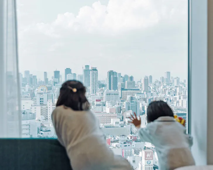Two children in bathrobes peer out the glass window at Osaka City at Centara Grand Hotel Osaka.