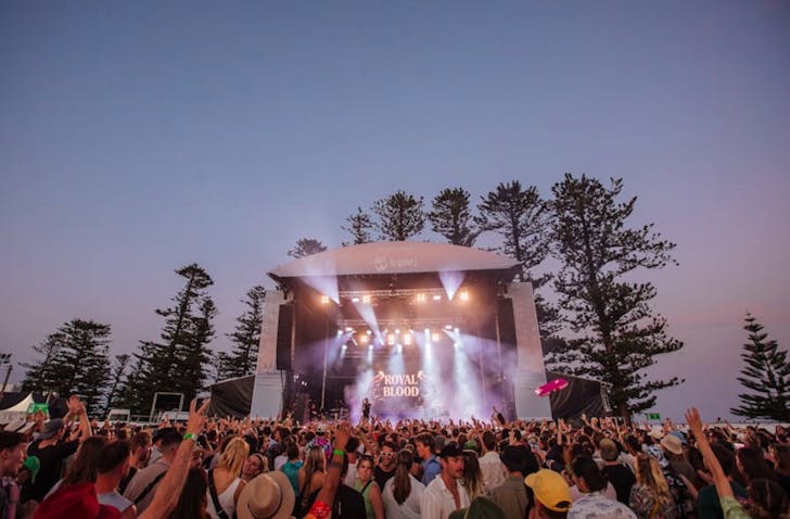 A wide shot of a festival crowd in front of a huge stage. The stage and festival crowd are outdoors, with Kiama beach trees in the background and a sunset sky. Colourful lights are coming from the stage and the crowd is dancing with their hands up.