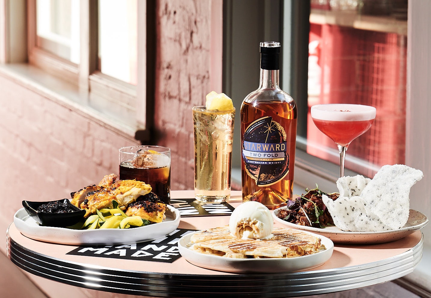 A suite of delicious eats sit on a pink table with pink neon signs lit behind it.