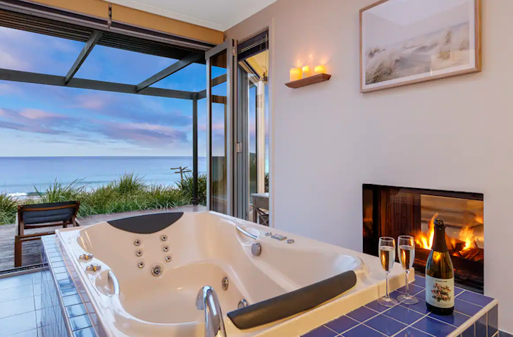 A rectangle-shaped spa looks out to a view of the ocean through an open floor-to-ceiling window, sitting next to a roaring fire place.