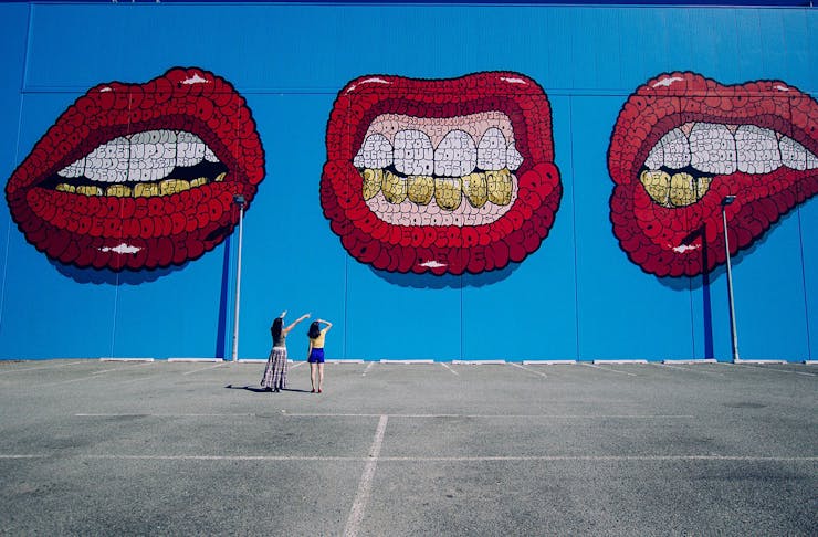 Two people gaze up at a huge street artwork showing three red mouths on a blue background.