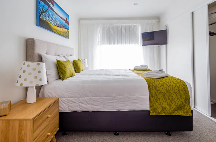 A large white bed with green cushions fits perfectly inside a small bedroom with a TV on the wall. 