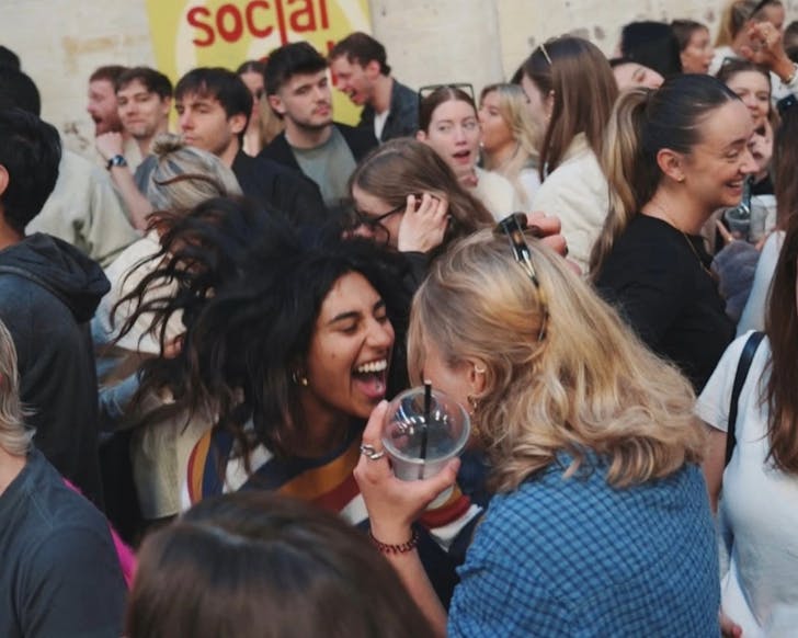 Two girls face each other in a calm crowd of dancers. They are singing at each other in a fun way, holding their coffees in hand. People are smiling and dancing around them, and the setting suggests daytime lighting.