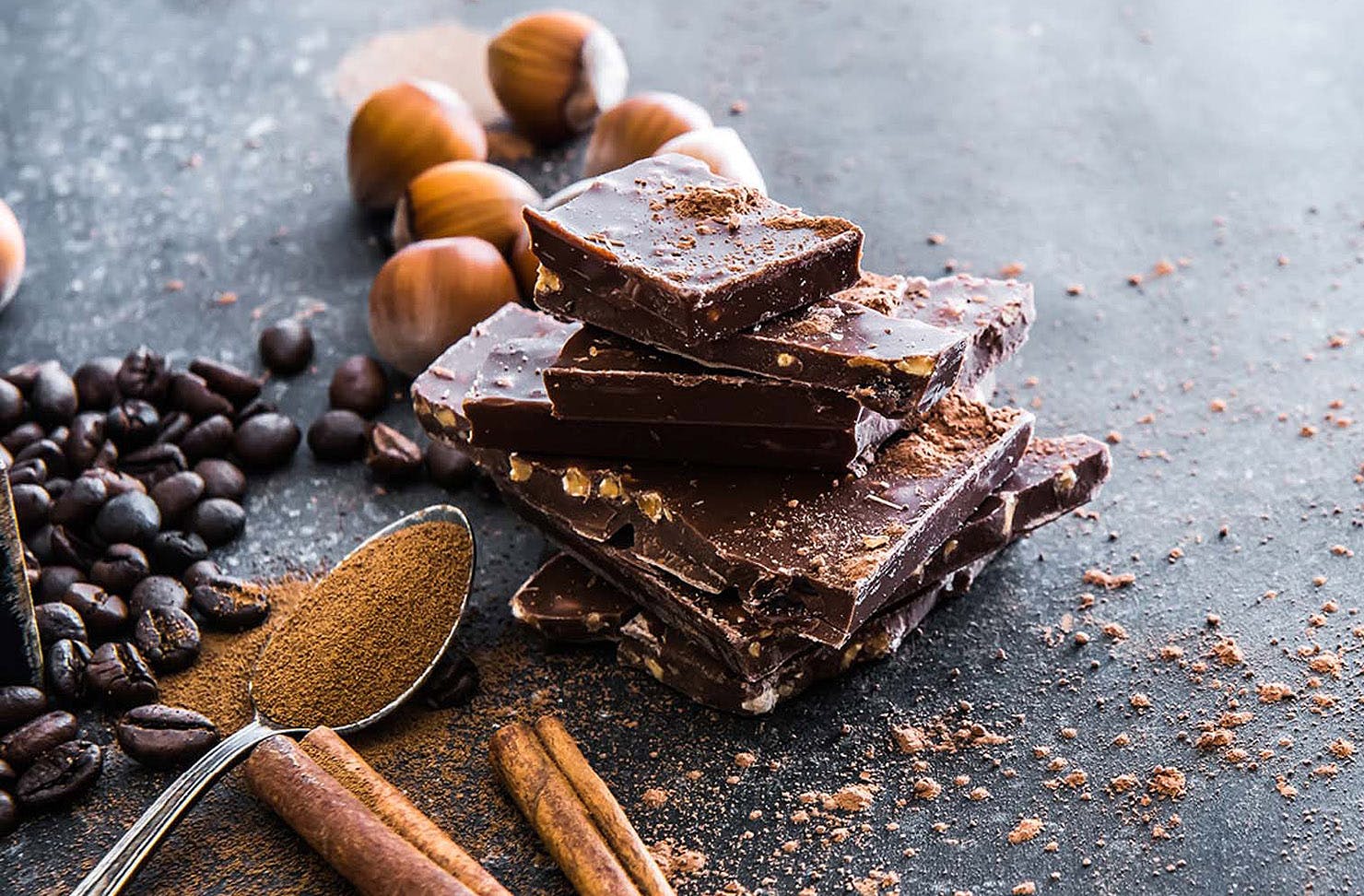 Broken up bars of chocolate rest on slate with a spoonful of cocoa powder and coffee beans and hazelnuts around.