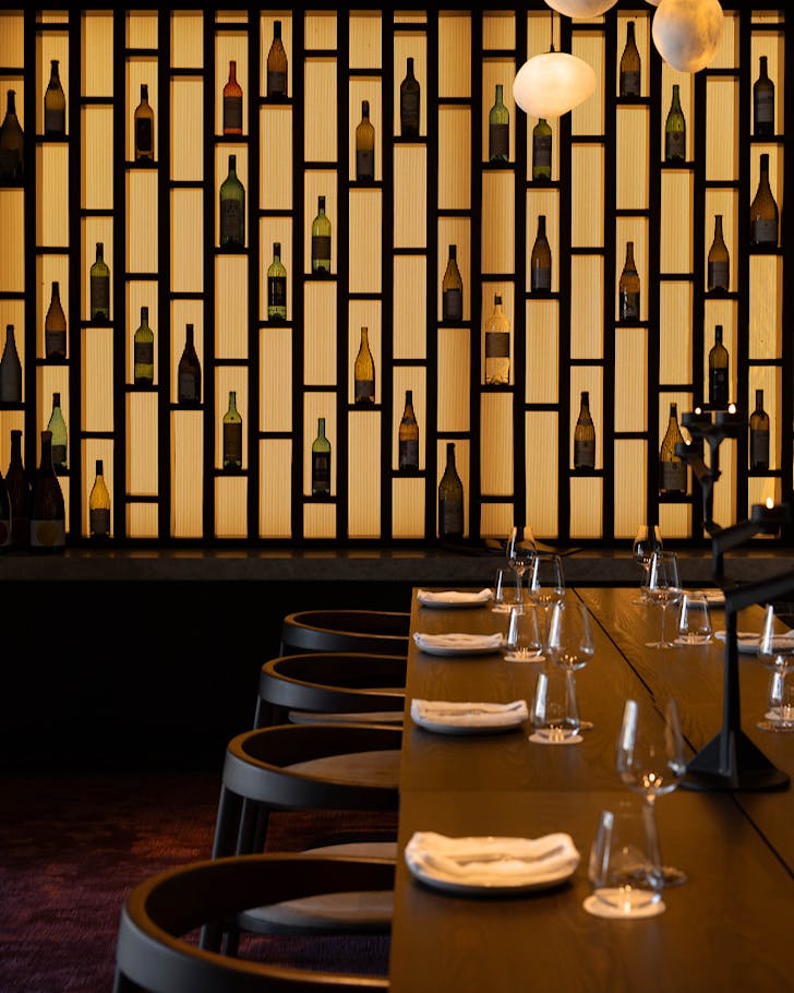 A dining table in front of a lit geometric wall studded with empty wine bottles.