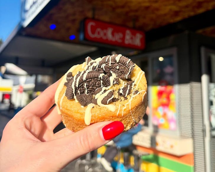 Someone with red nail polish holds up a loaded cookie in front of the Cookie Bar shopfront. 
