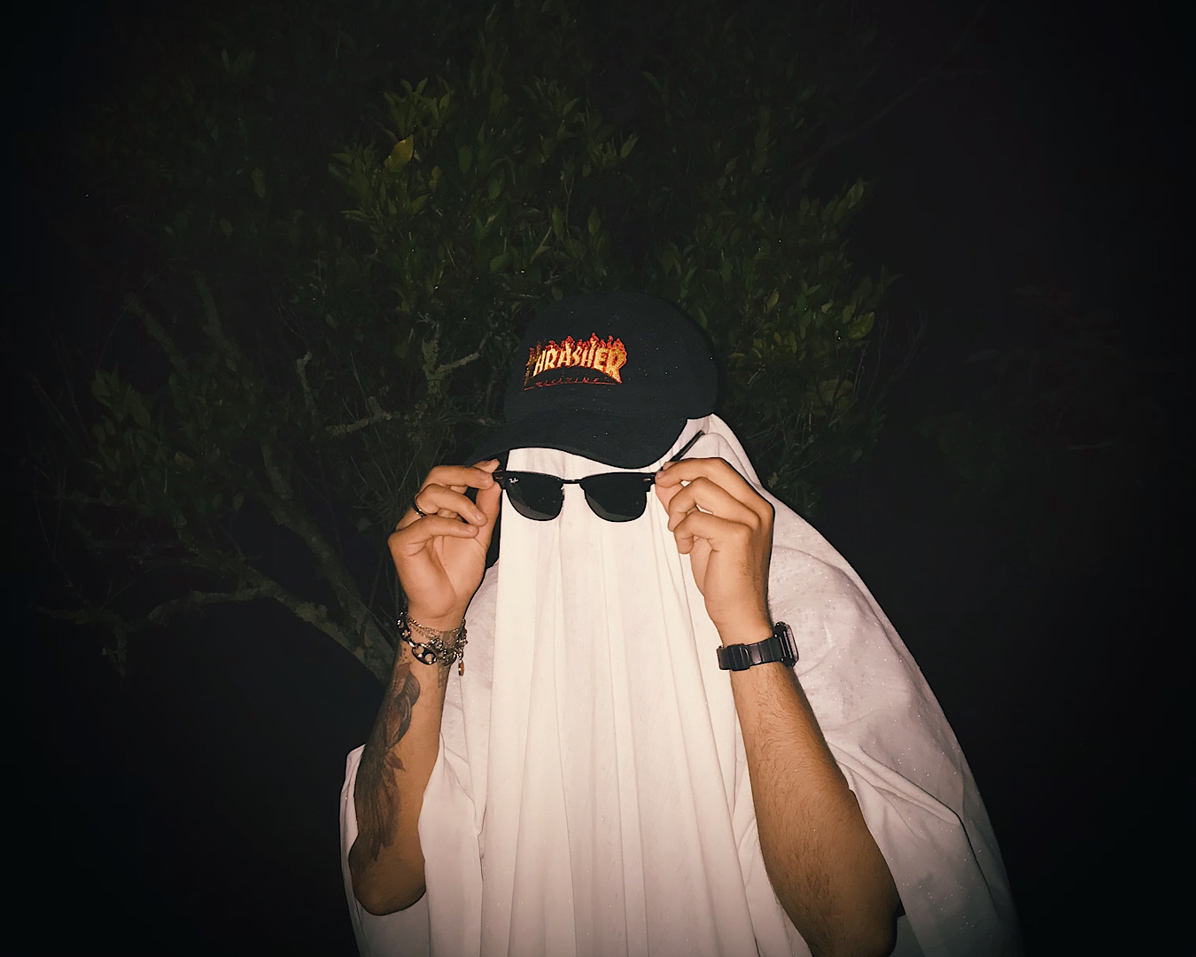 A person slides on a pair of Ray Ban sunnies to complete their ghost Halloween costume of a white sheet, a ‘Thrasher’ cap, a watch and a bracelet. We glimpse an impressive tattoo on their left forearm. 