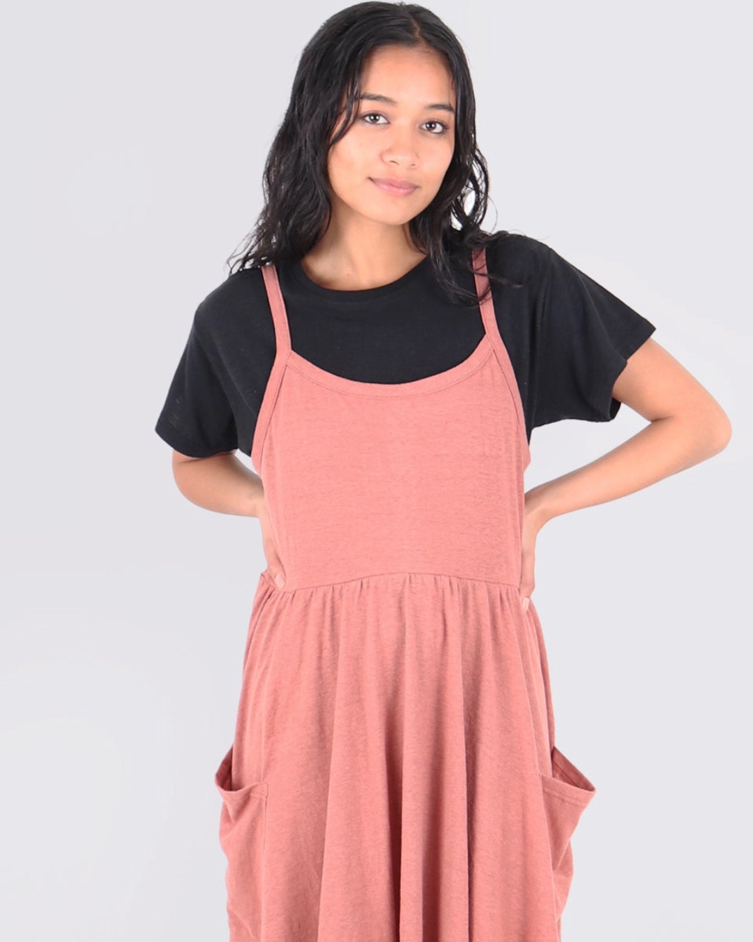 A woman wears a salmon babydoll dress with pockets over a black t-shirt. 