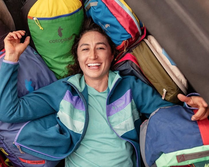 A woman lies on top of a bunch of colourful Cotopaxi bags. 