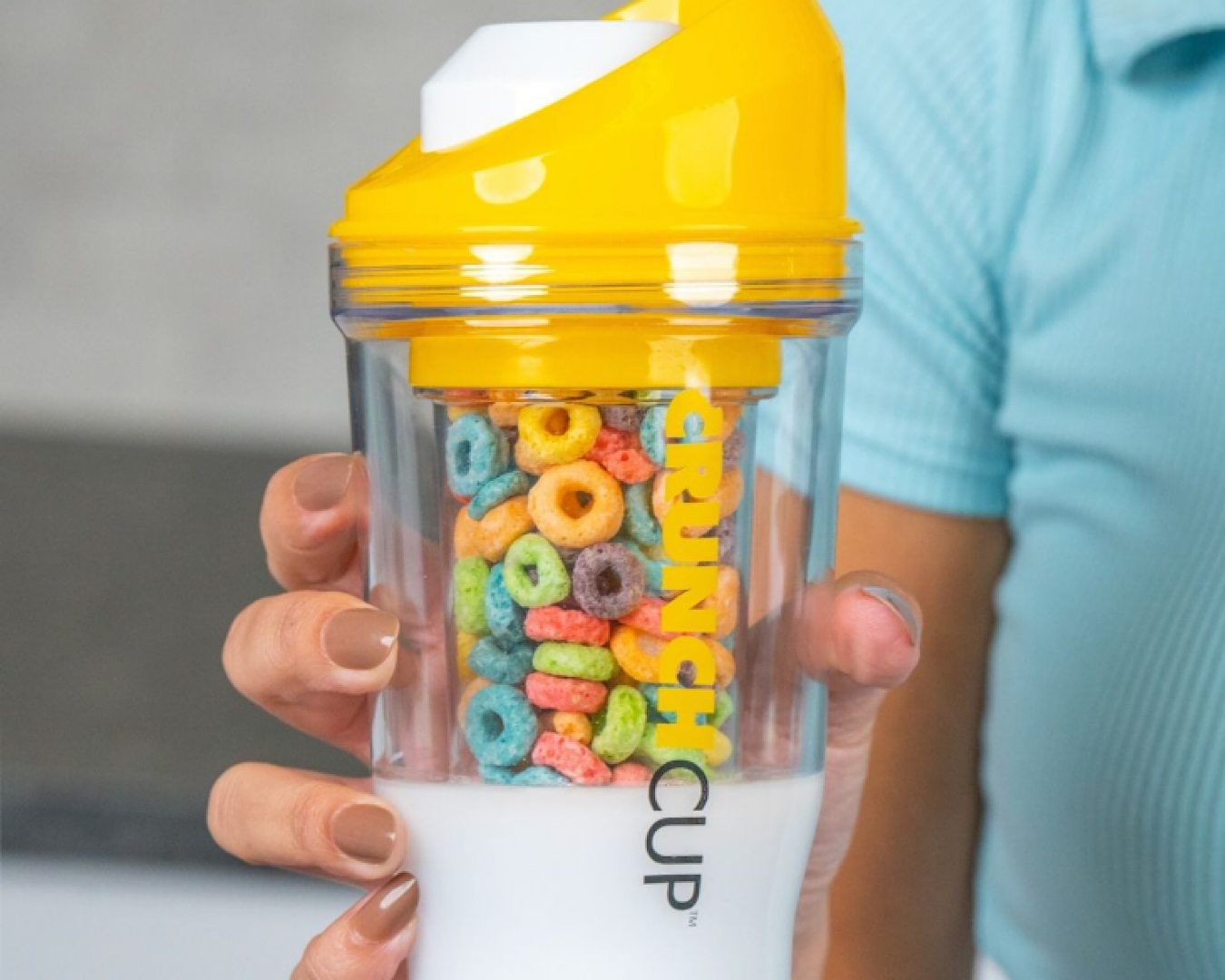 A hand holds a reusable plastic cup filled with milk and brightly coloured cereal. 