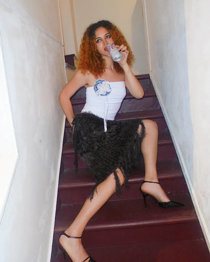 A woman sits on a flight of burgundy stairs decked out in vintage and sipping on a silver can of Coca Cola.