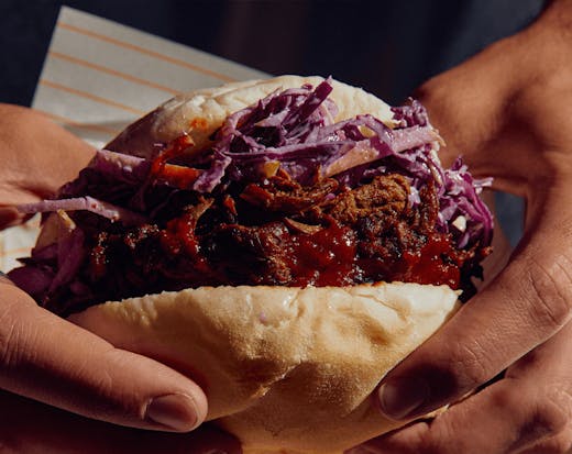 Two hands holding up a roll filled with red cabbage and pulled pork. 
