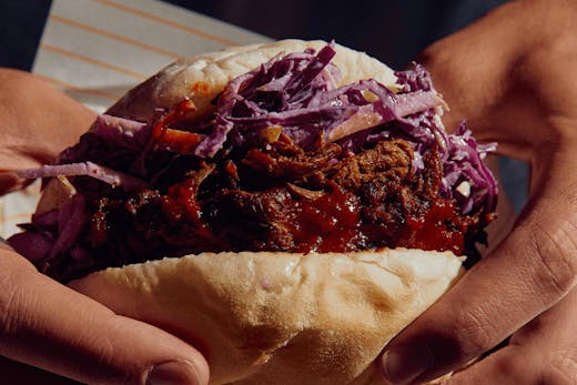 Two hands holding up a roll filled with red cabbage and pulled pork.