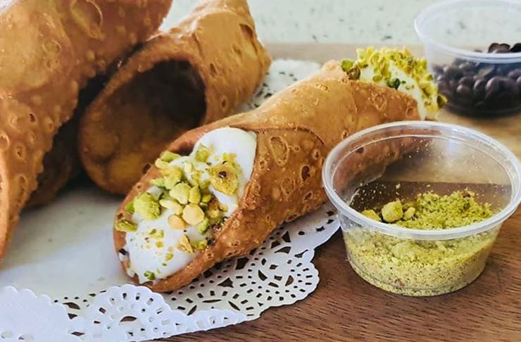 A DIY cannoli with a fresh ricotta and pistachio filling sits on a timber chopping board with leftover crushed pistachios to the side in a small plastic dish.