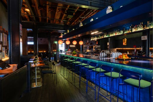 A mid shot of the interior of the cocktail bar room, where the room is low lit with a sleek bar design that is illuminated with neon blue lights underneath the bar table. Many seats line the bar and there are lower, more formal tables available.