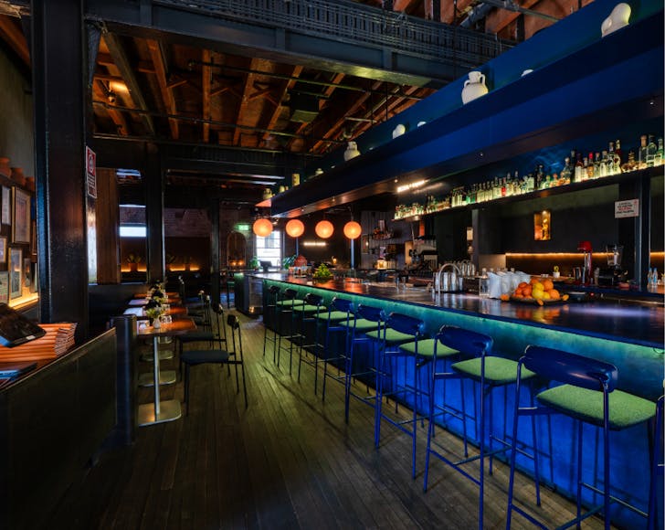 A wide shot of the cocktail bar that features dark design colours with pops of blue and orange neon lighting elegantly emitting from beneath and around the bar table. Stools along the bar are lined up as well as standard 2-person seating spots to the left. 