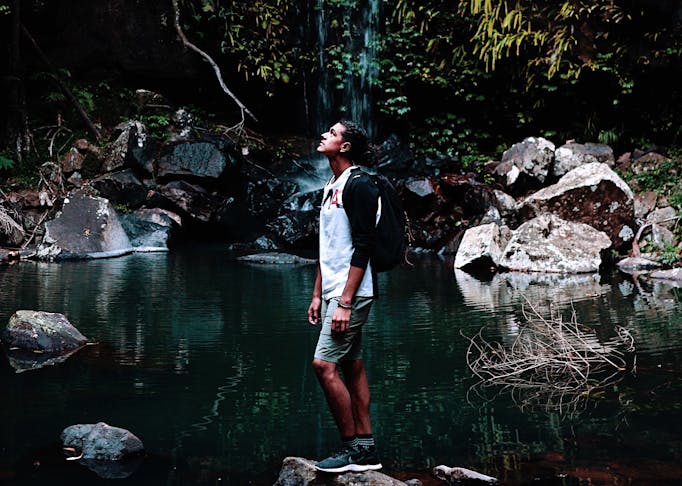 A man stands on a rock in front of a waterfall looking up towards the sky.