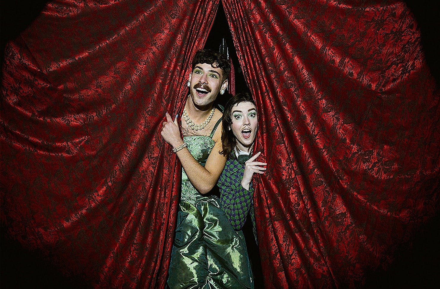 Two cabaret performers look out at the crowd from red velvet curtains. 