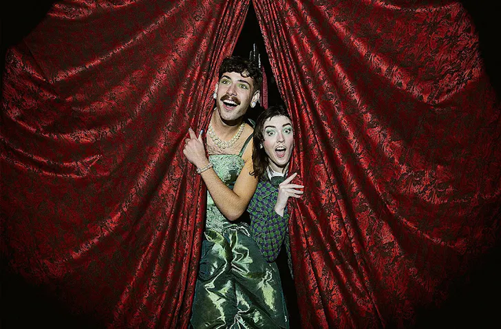 Two cabaret performers look out at the crowd from red velvet curtains. 