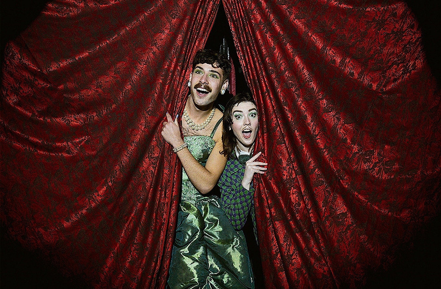 Two cabaret performers look out at the crowd from red velvet curtains. 