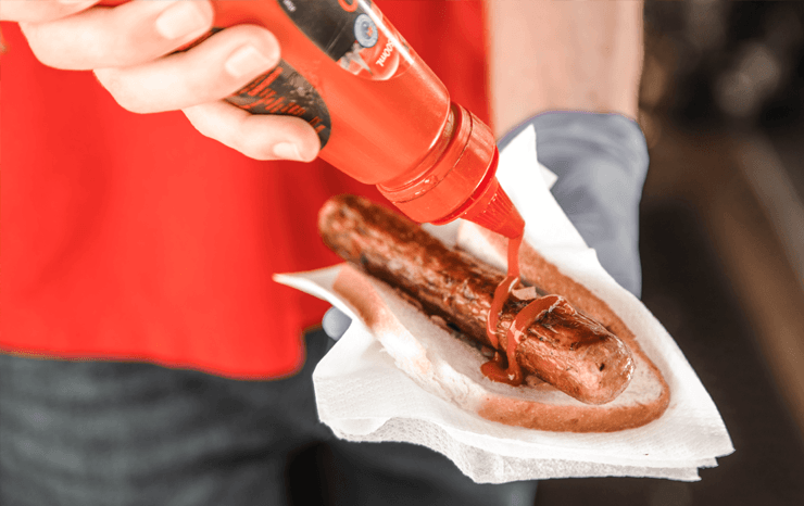 A person putting sauce on a sausage.