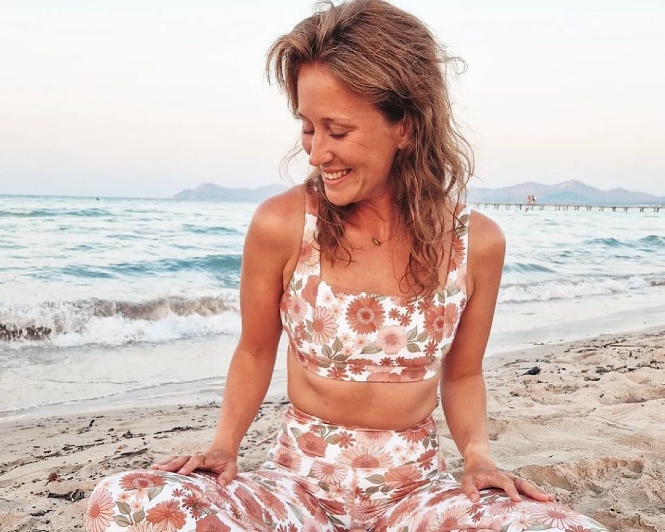 A woman sits cross legged on a beach wearing Dharma Bums leggings and a crop top. She looks off to one knee and smiles. 