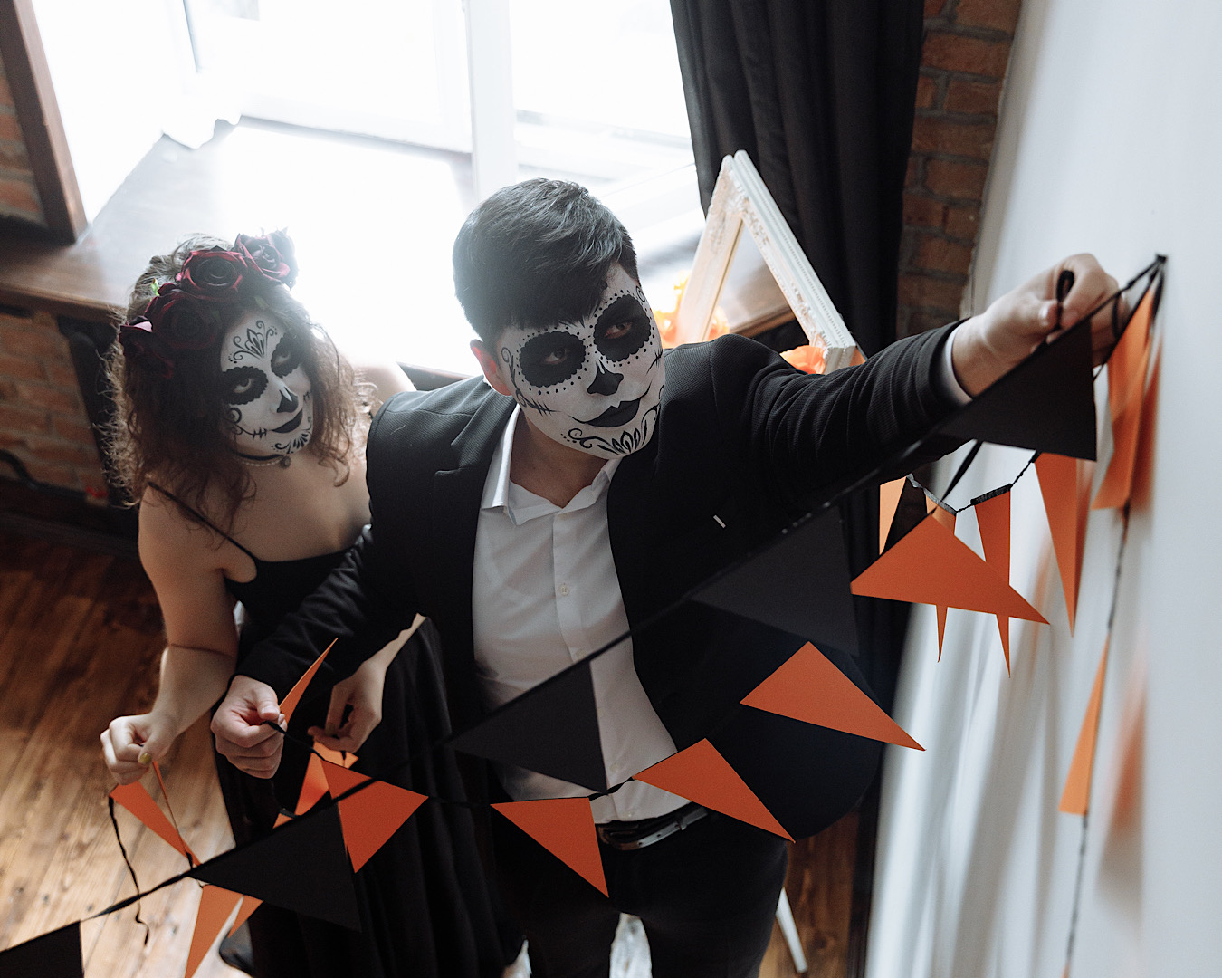 Two people dressed in Dia de Los Muertos-style skeleton Halloween costumes put up an orange and black bunting. 