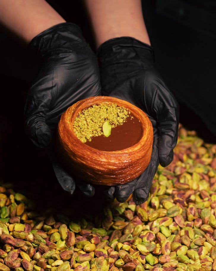 Gloved hands holding Dipcro pistachio Dubai chocolate tart above layers of pistachios