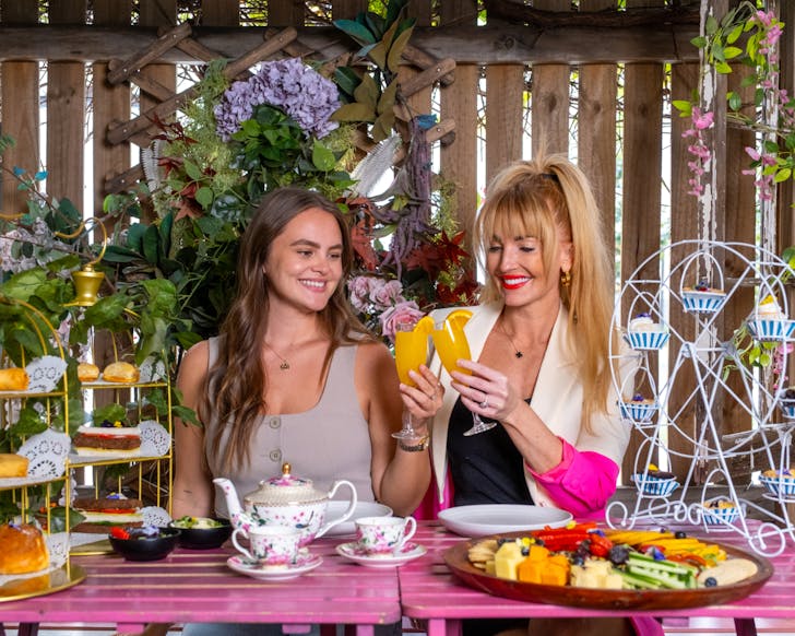 Two women sit smiling outdoors at a bright pink table. The table is covered in pretty plates of food including decorative cupcakes and trays of pastries. The two women are smiling while holding their glasses of orange drink. 