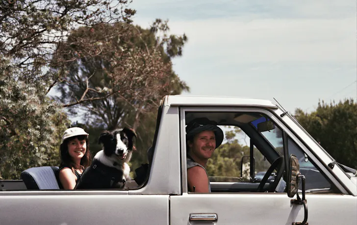 A ute with a woman and border collie in the back and a man driving smiling. 