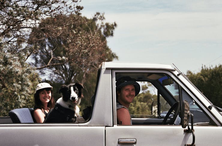 A ute with a woman and border collie in the back and a man driving smiling. 