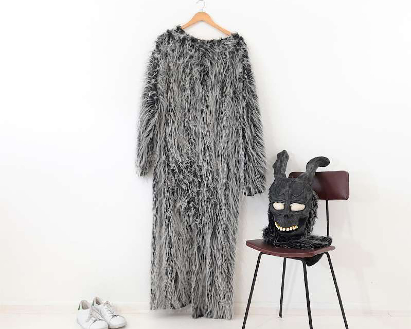 A fluffy black and white horror rabbit gown hangs on a coat hanger beside a freaky rabbit mask on a brown chair and a pair of white sneakers. Together, they form a Donny Darko costume by Desiree Costume Hire.  
