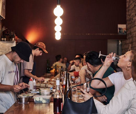 A bar with people drinking on one side and people preparing food on the other. 