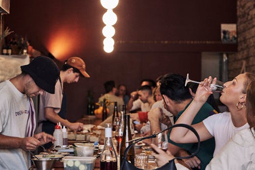 A bar with people drinking on one side and people preparing food on the other.