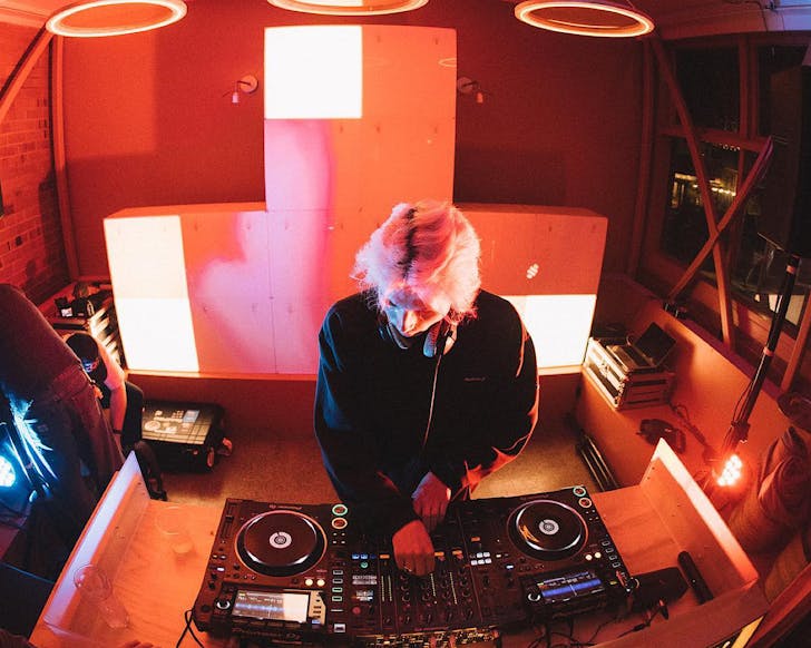 Birdâs eye view of a pink-haired DJ spinning some tracks at EAST ST. HALL bathed in a pinky coral light. 